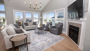 A spacious living room with a fireplace, large windows, and a chandelier. - Oceanside, 8 BRs, Private Pool, Theater Room, Rec Rm, Hot Tub, OL126, Knot Tide Up (Corolla)