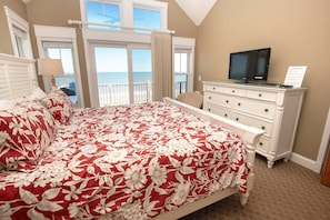Quarto com cama grande, televisão sobre cômoda e vista para o oceano pelas janelas.