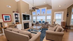 A spacious living room with beige sofas, a fireplace, a TV, and a view of the ocean through large windows. - OCEANFRONT, 10 BRS, ELEV, Pool, Rec Rm, Theater Rm, OH1251, Beach Plaza (Corolla)
