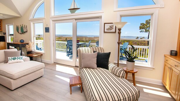 A spacious room with a large window overlooking a deck, a sofa, and a small table with a lamp. - Soundfront, ELEV, Private Pool, Pool Table, Water Views, VOH1, Soundly Sea-Cluded (Corolla)