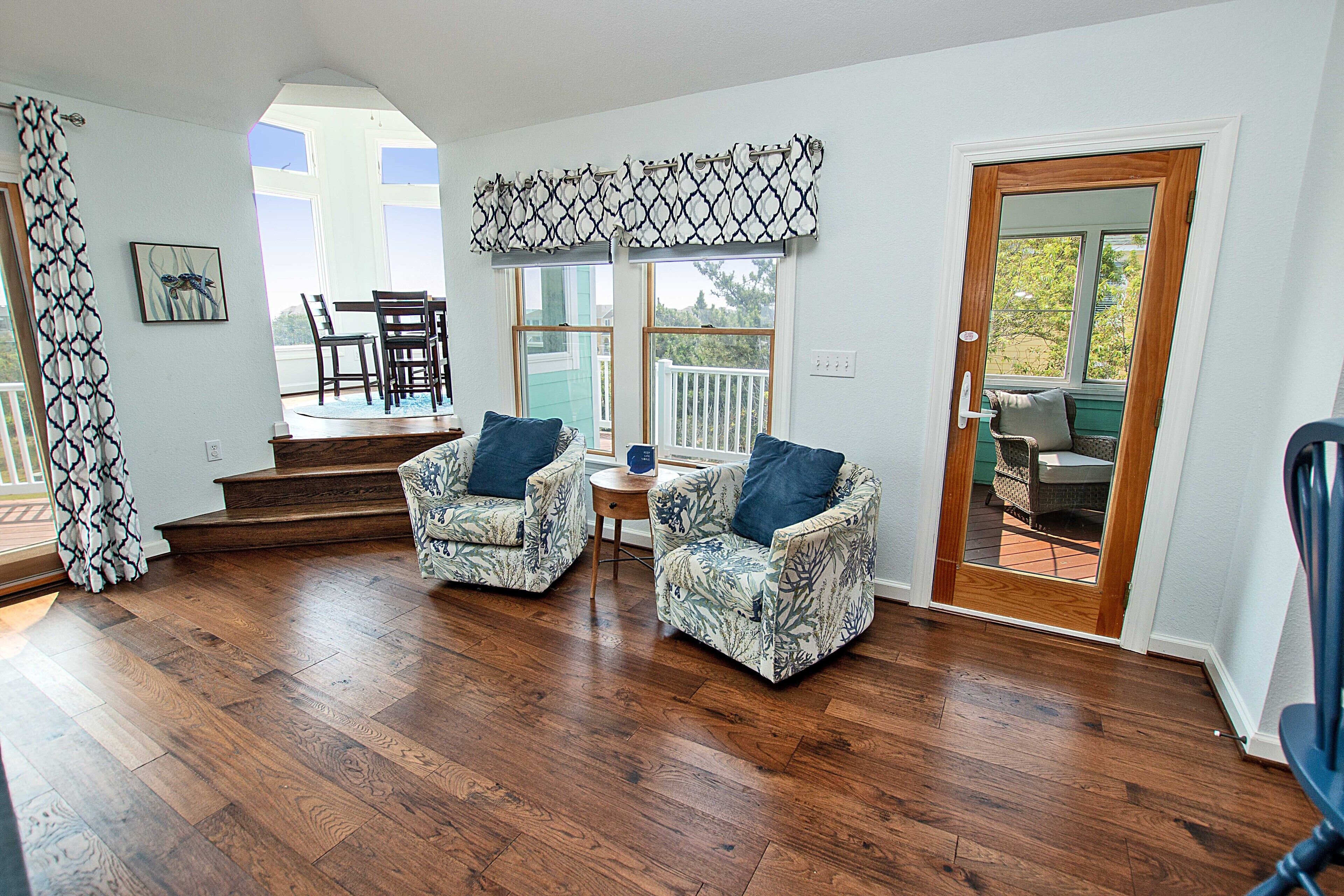 A spacious living area with hardwood flooring, two patterned armchairs, a dining table with chairs, and a large window with a view of the outdoors.