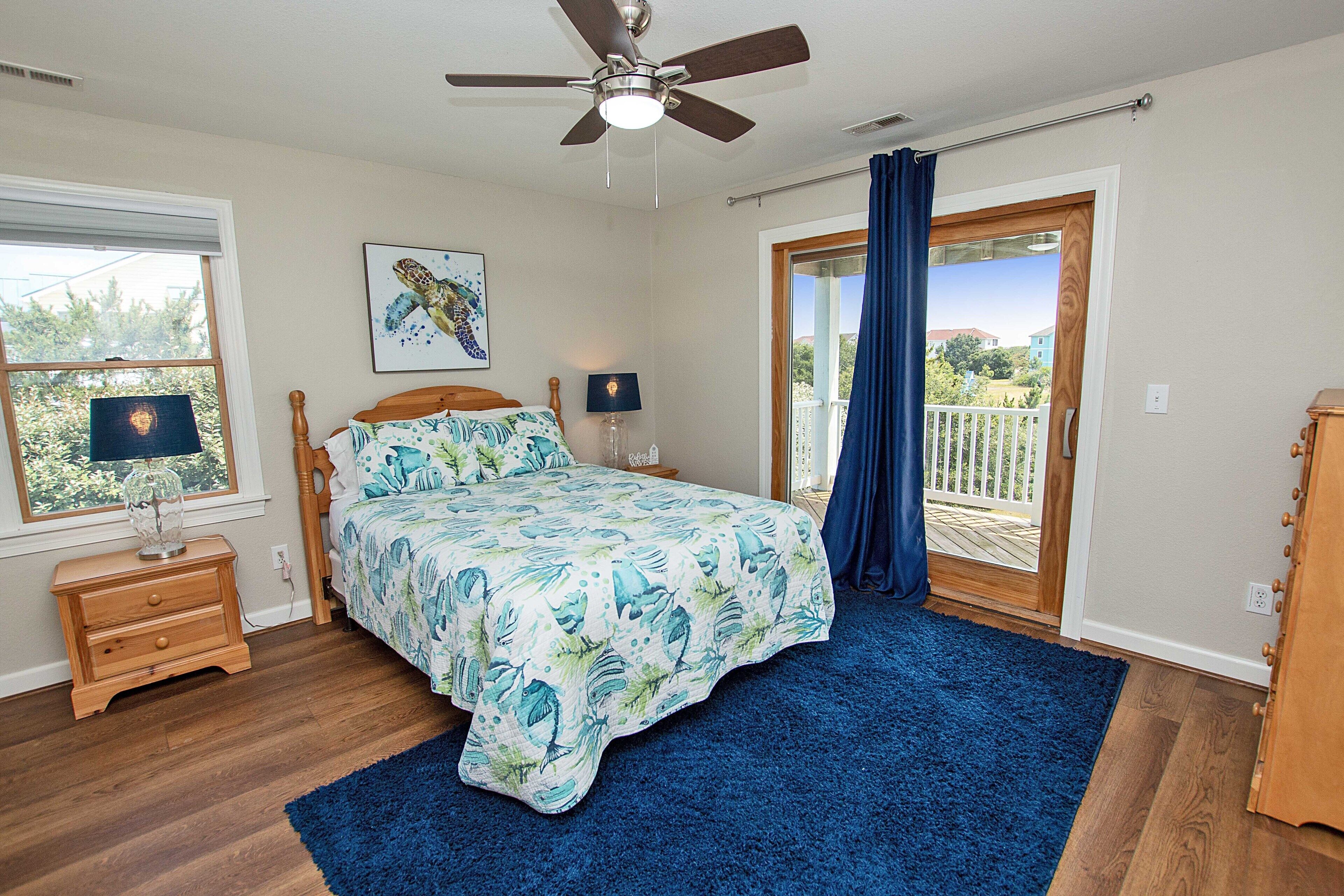 A bedroom with a bed, two bedside tables, a ceiling fan, a window with curtains, and a view of the outdoors.