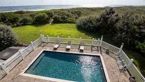 Ein rechteckiger Pool mit weißem Zaun und Liegestühlen, umgeben von einer Rasenfläche und dichter Bepflanzung, mit Blick auf eine Küstenlandschaft.