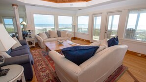 A spacious living room with a large window overlooking the sea, a cozy sofa set, and a wooden coffee table. - OCEANFRONT, SUN-SUN, Pool, ELEV, Ocean Vws, PM100, Ships In (Duck)