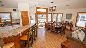 A spacious kitchen with a central island, wooden dining table, and chairs. There is a balcony with a view, and the room has wooden flooring and white walls. - Oceanside, Private Pool, Hot Tub, Close to Beach Access, OL656, Bali Dreams (Corolla)