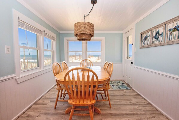 A dining area with a wooden table and chairs, a hanging light fixture, and a view of the beach through large windows. - OCEANFRONT, Ocean views, Dogs Welcome! KH10, Beach House (Kitty Hawk)