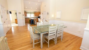 A dining area with a glass table and white chairs, wooden flooring, and a kitchen in the background. - Semi-Oceanfront, Private Pool, Hot Tub, Game Room, KD1502, Tide Watcher (Kill Devil Hills)