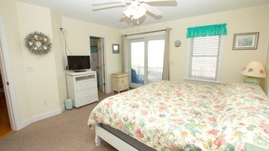 Une chambre à coucher avec un lit, un téléviseur posé sur une commode, un ventilateur de plafond, une fenêtre avec des stores et une porte donnant sur un balcon.