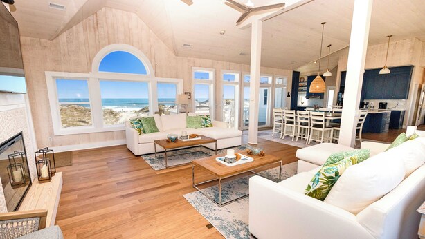 Un vaste espace de vie avec une grande fenêtre donnant sur la plage, un canapé blanc, une table basse et un coin cuisine avec un ensemble de salle à manger.