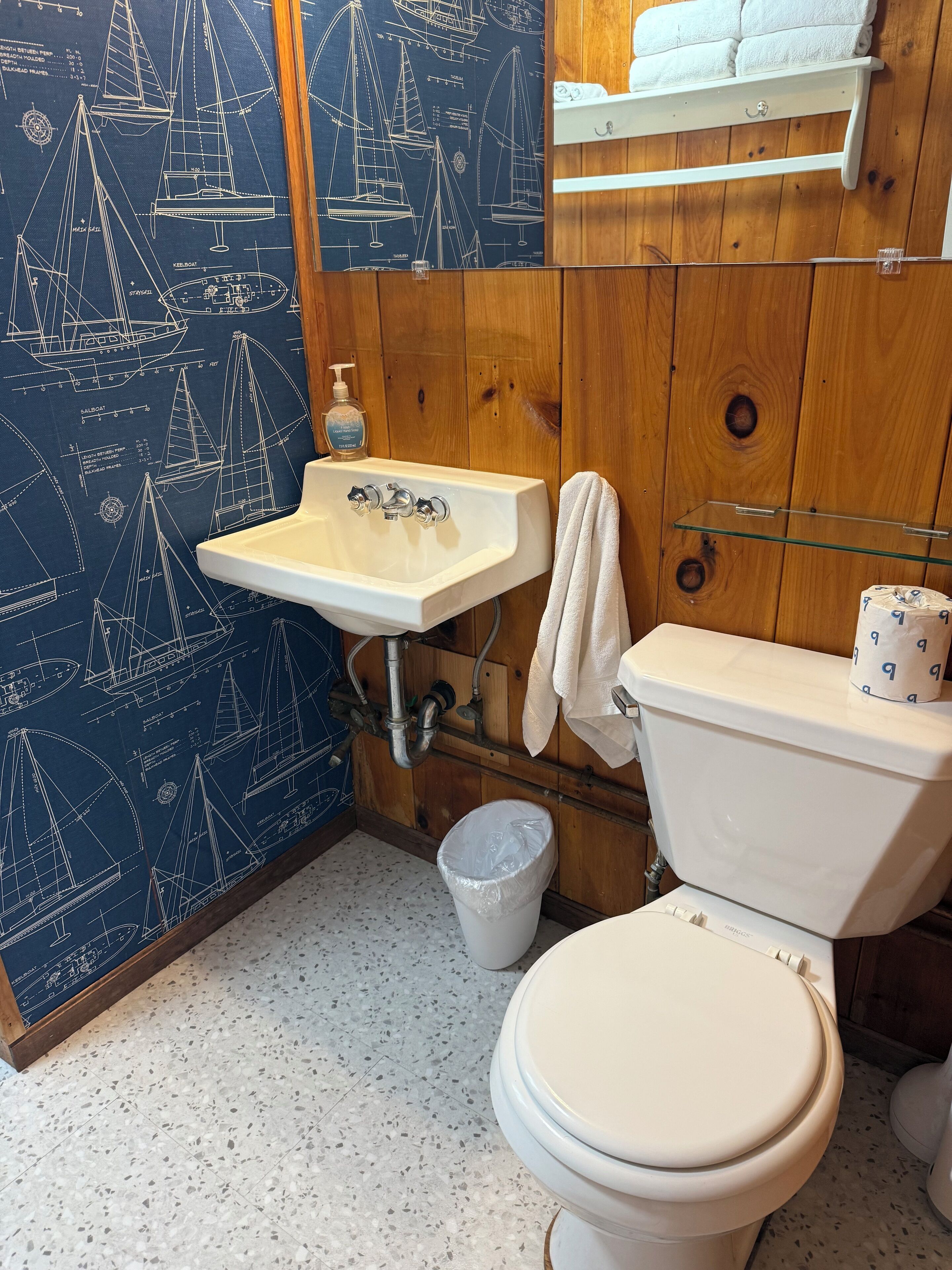 Une salle de bain avec un papier peint sur le thème de la marine, un lavabo blanc, des toilettes et une petite poubelle.