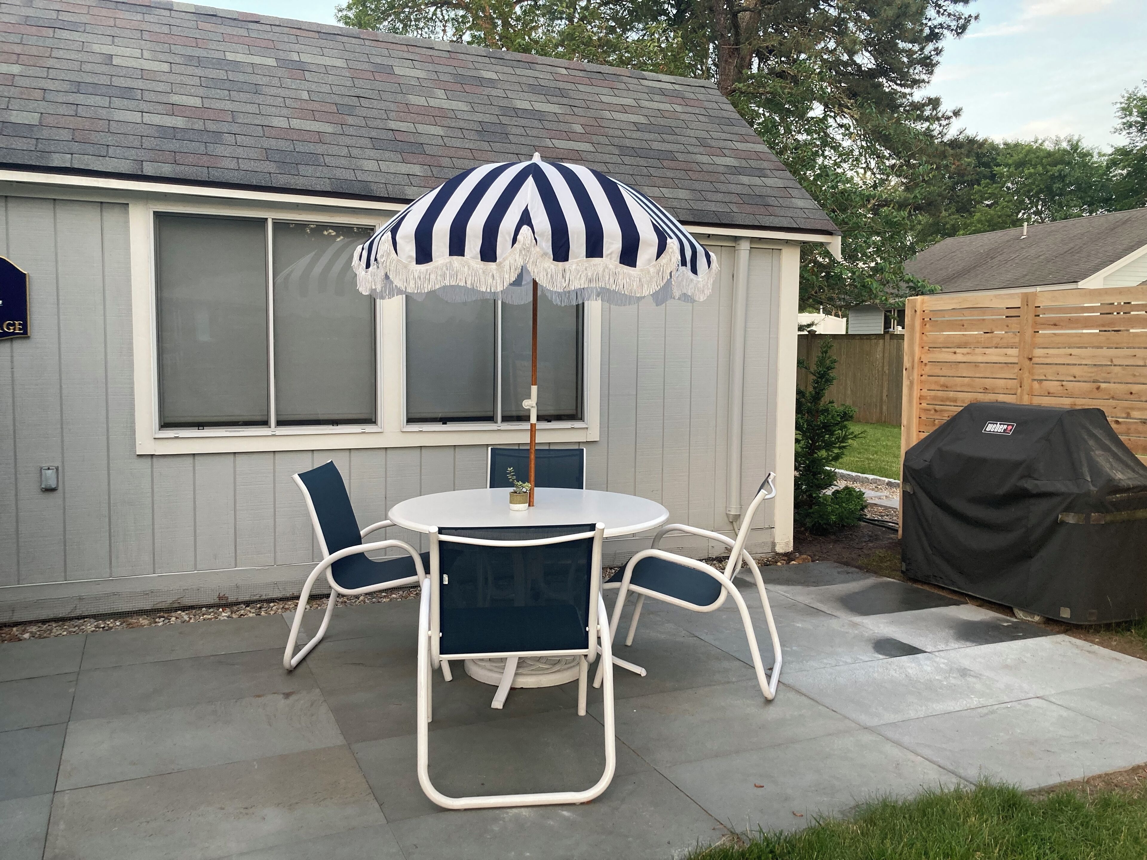 Eine Terrasse mit rundem Tisch und Stühlen, einem Streifen-Sonnenschirm und einem Grill.