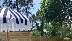 A patio with a round table and chairs, a striped umbrella, and a wooden fence. -  Steps to private Beach, Golf Course, AC, tandem Kayak for Guests (South Yarmouth)