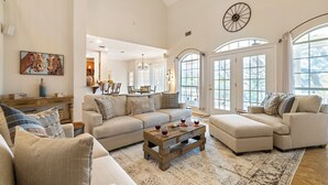 Un salón amplio con un sofá seccional grande, una mesa de centro y una chimenea. Hay ventanas grandes y vistas al exterior.