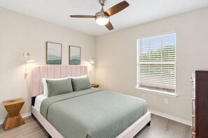 Un dormitorio con cama, ventilador de techo, luces fijadas en la pared, una ventana con persianas y una mesita de noche.