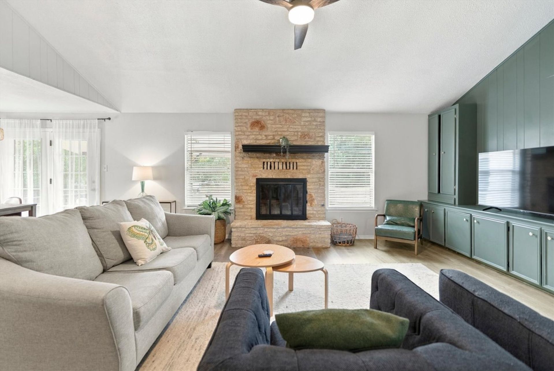 Relax in this inviting Austin living room with a cozy sectional, a charming stone fireplace, sage-green cabinetry, and a large TV. Natural light and warm accents create a welcoming space to unwind after exploring the city.