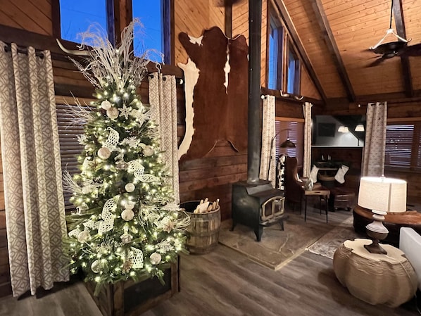 A rustic cabin interior with a decorated Christmas tree, wooden walls, a stone stove, and a dining area.