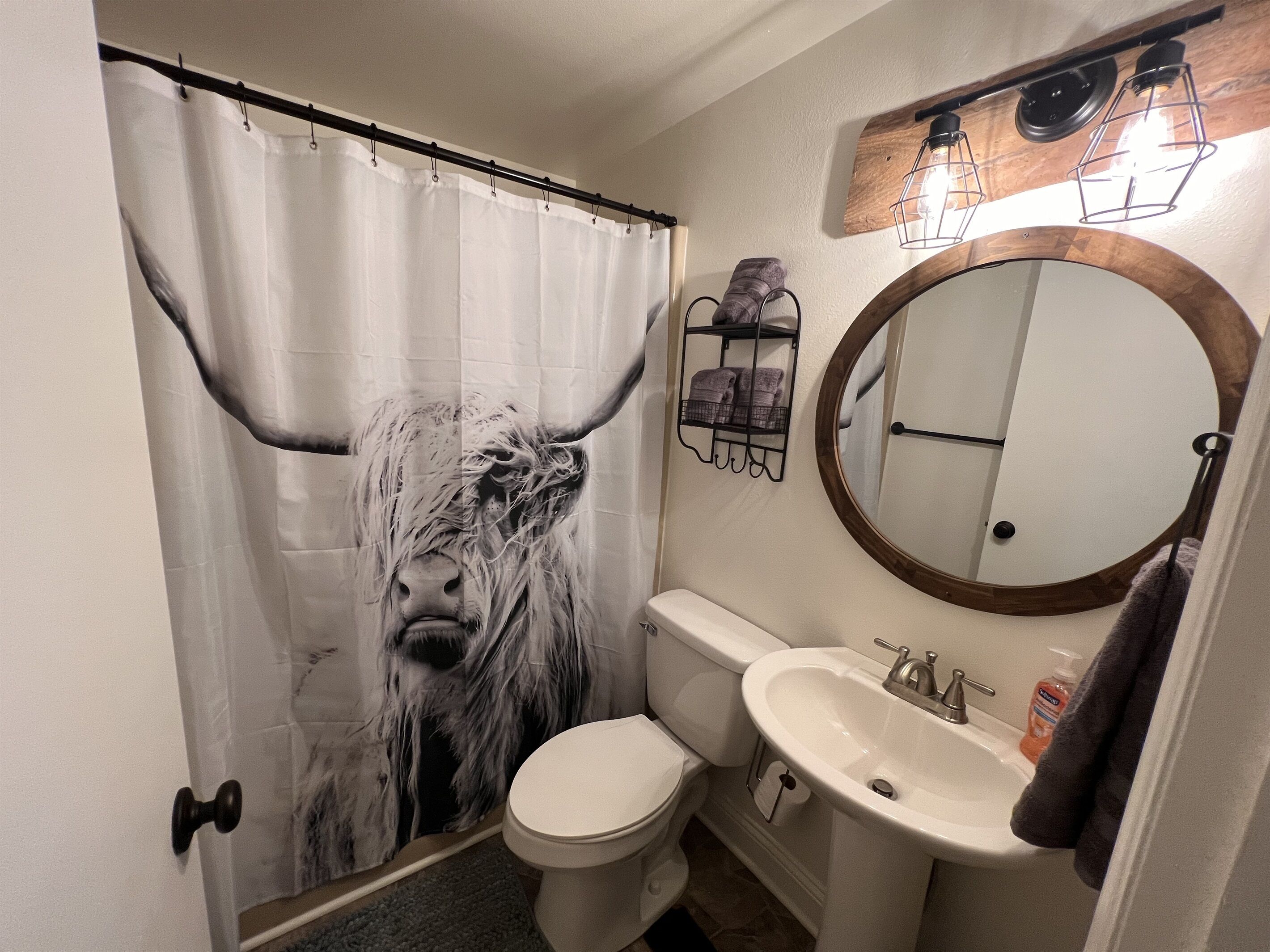 Ett badrum med en duschdraperi som har ett komotiv av en kos ansikte, en toalett, ett handfat med spegel och en handduksställning.