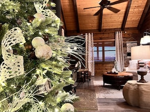 A cozy log cabin interior with a decorated Christmas tree, wooden furniture, and a ceiling fan.