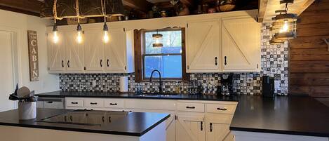 Una cocina con armarios blancos, encimera negra y ventana. Hay luces colgantes y un letrero que dice "CABIN".