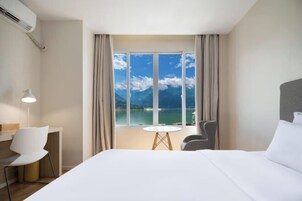 A hotel room with a large bed, a desk with a chair, a small table, and a view of a body of water and mountains.
