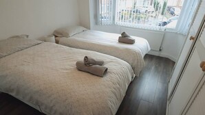 A hotel room with two beds, each with a rolled towel on top. The room has a window with blinds, a radiator, and a wooden floor. - Beautiful modern 3-Bed House in Birmingham (Birmingham)