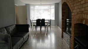 A living room with a sofa, a dining table with chairs, a fireplace, and a large window with blinds. - Beautiful modern 3-Bed House in Birmingham (Birmingham)