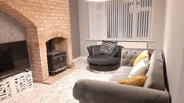 A living room with a brick fireplace, a grey sofa with yellow cushions, a black armchair, and a white radiator above the sofa. - Beautiful modern 3-Bed House in Birmingham (Birmingham)