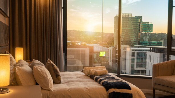 A hotel room with a large window, a bed with pillows, a lamp, and a view of the cityscape. - Black Marble - Menlyn Residence (Pretoria)
