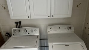 Une buanderie équipée de deux machines à laver blanches et d’un sèche-linge, avec un meuble blanc au-dessus et un porte-serviettes fixé au mur.