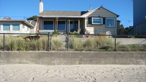 Een huis aan het strand met een omheinde tuin, een betonnen barrière en een zandstrand op de voorgrond.