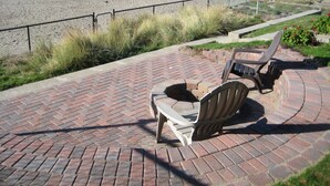 Een pad van bakstenen leidt naar een strand, met twee Adirondack-stoelen en een metalen hek.