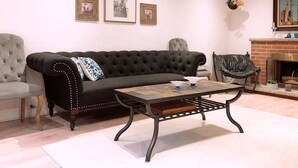Una sala de estar con un sofá negro, una mesa de centro de madera, un sillón gris y una chimenea con un cuadro colgado encima.