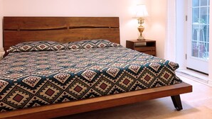 Una cama con colcha estampada, cabecera de madera, mesita de noche con lámpara y vista al exterior a través de una ventana.