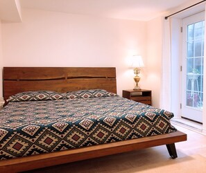 A bed with a patterned bedspread, a wooden headboard, a bedside table with a lamp, and a view of the outdoors through a window.
