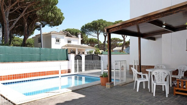 A swimming pool with a tiled edge, a covered patio area with white plastic chairs and tables, and a white building with a tiled roof in the background. - Chalet 'Poniente' with Pool and Garden (Roche)