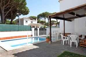 A swimming pool with a tiled edge, a covered patio area with white plastic chairs and tables, and a white building with a tiled roof in the background. - Chalet 'Poniente' with Pool and Garden (Roche)