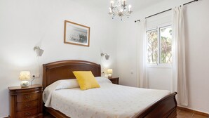 A bedroom with a wooden bed, a nightstand with a lamp, a wall-mounted picture, and a window with curtains. - Chalet 'Poniente' with Pool and Garden (Roche)