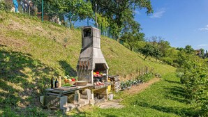 A stone barbecue grill with a table set up nearby, surrounded by a grassy area and trees.