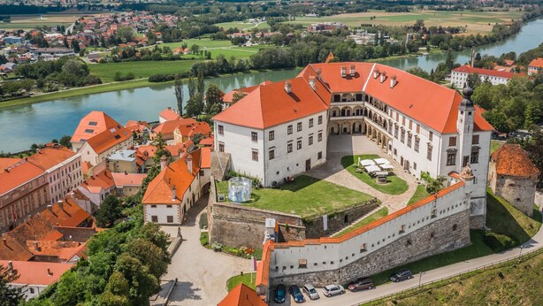 A historic building with a courtyard, surrounded by a fortified wall, overlooking a river and a town.