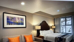 Une chambre d’hôtel avec un lit, un canapé gris agrémenté de coussins orange, une table de chevet avec des lampes et une fenêtre donnant sur un paysage enneigé.
