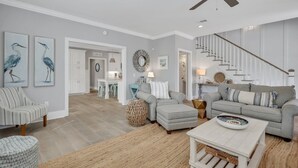 A spacious living room with a grey sofa, a striped armchair, and a coffee table. A staircase with a wooden railing leads to another level. - Amazing Island Oasis, Heated Pool, Outdoor Bar Area with TV, Bocce Ball Court & Putting Green (Saint Simons Island)