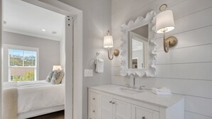 A modern bathroom with a white vanity, a large mirror, and a wall-mounted light fixture. - Amazing Island Oasis, Heated Pool, Outdoor Bar Area with TV, Bocce Ball Court & Putting Green (Saint Simons Island)