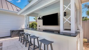 An outdoor bar area with a flat-screen TV, white wooden walls, and four bar stools. - Amazing Island Oasis, Heated Pool, Outdoor Bar Area with TV, Bocce Ball Court & Putting Green (Saint Simons Island)