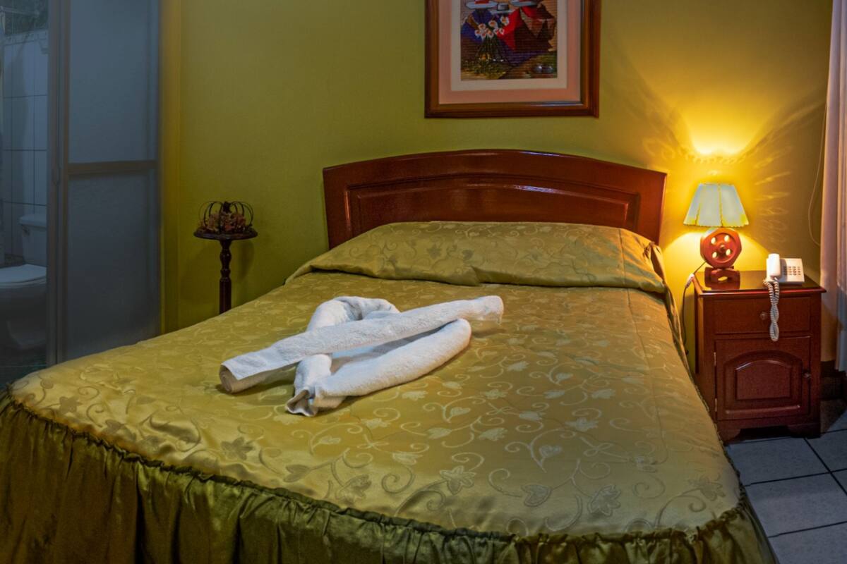Une chambre d’hôtel avec un lit, une table de chevet avec une lampe, un tableau encadré au mur et une salle de bain visible au fond.