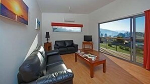 Un salon moderne avec un canapé en cuir noir, une table basse en bois et un téléviseur posé sur un meuble. Une grande fenêtre offre une vue sur le paysage urbain et les montagnes.