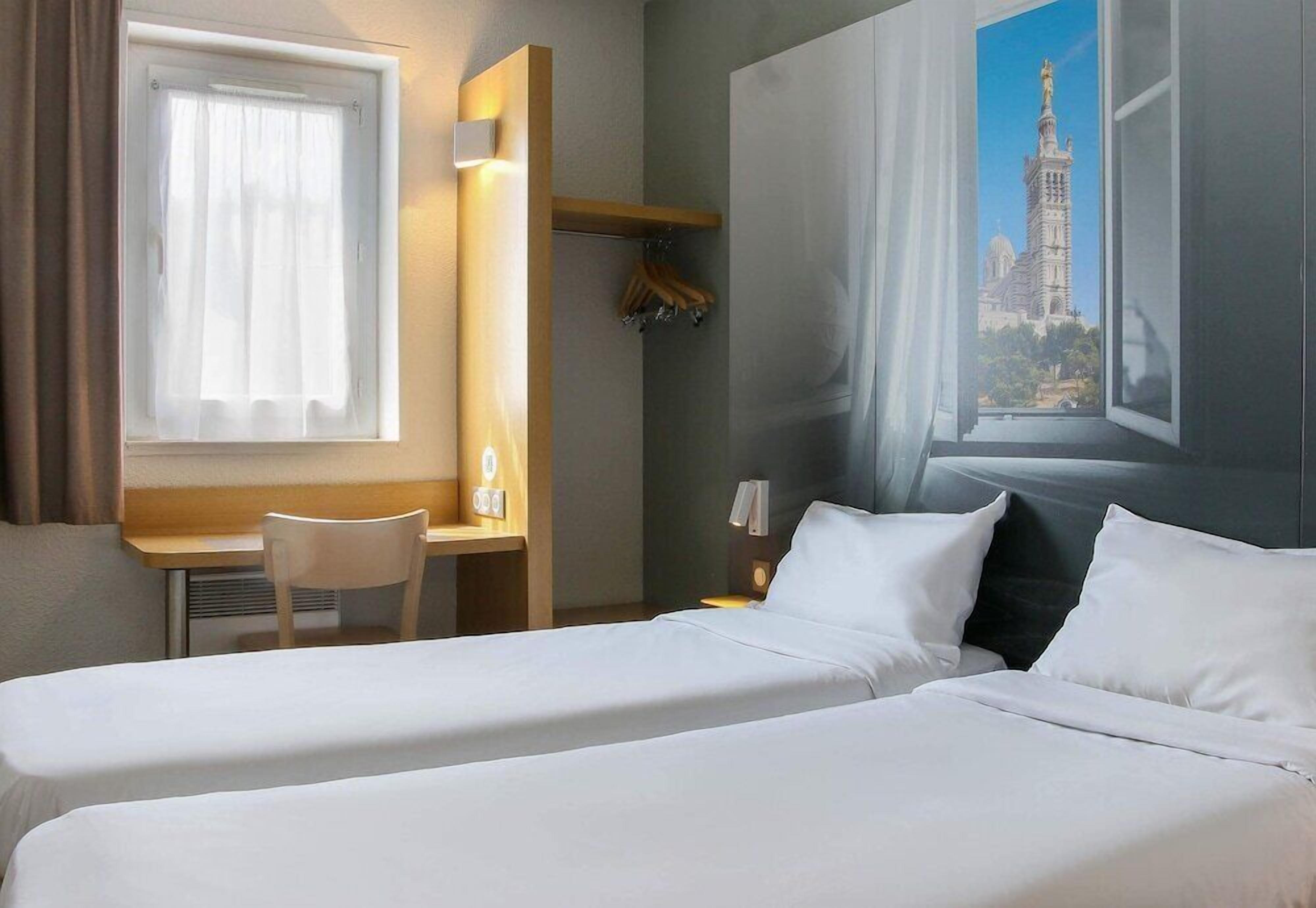 A hotel room with two beds, a desk, a chair, a window with a view of a building, and a wall-mounted shelf.
