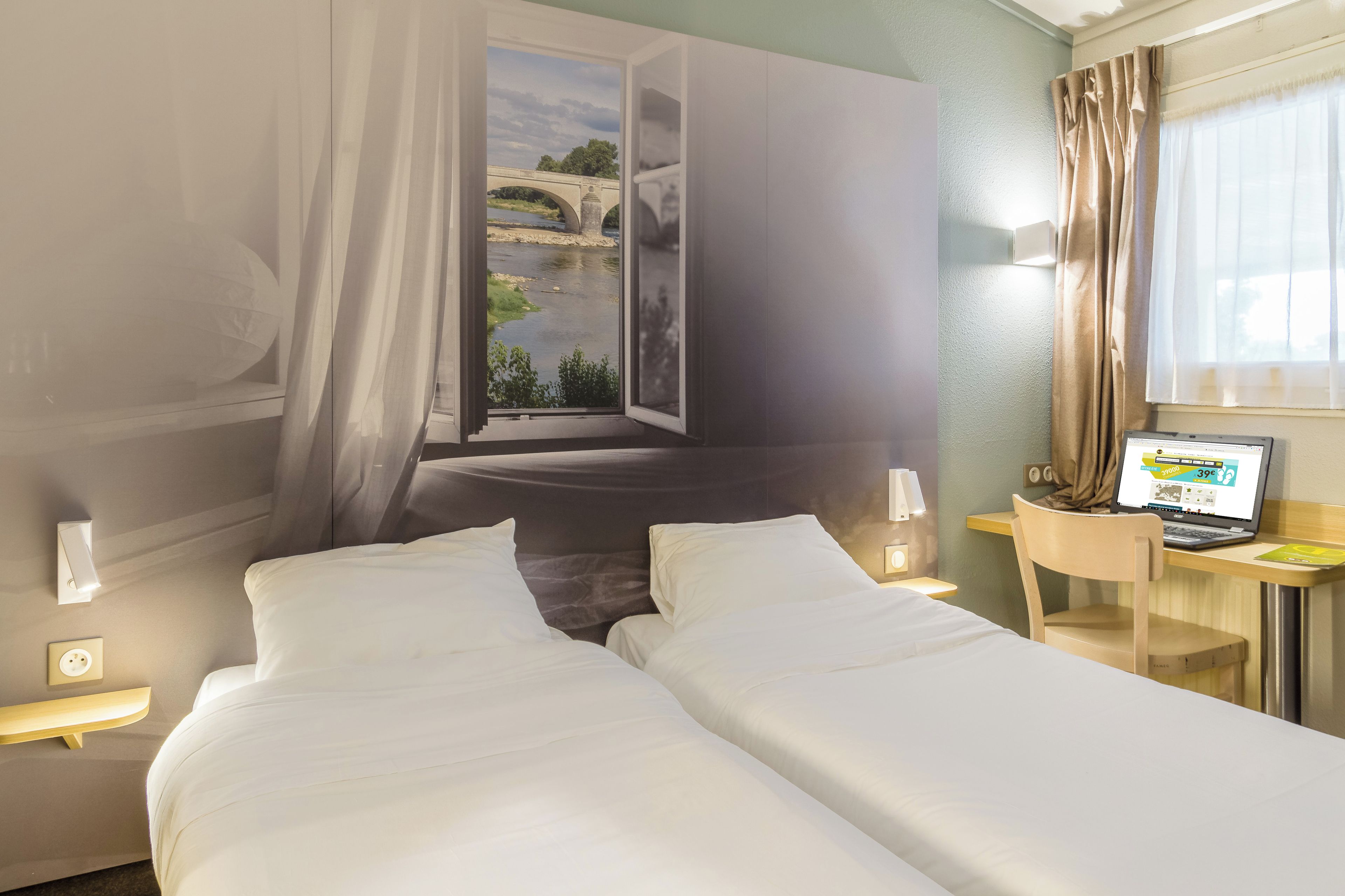 Hotellihuone, jossa on kaksi sänkyä, työpöytä kannettavalla tietokoneella, ikkuna, josta on näkymä sillalle, ja verho.