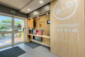 Un espace d’accueil comprenant un comptoir de réception en bois, un écran numérique et une porte vitrée donnant sur l’extérieur.