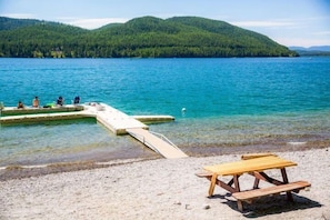 A wooden dock extending into a clear lake, surrounded by a sandy beach with a picnic table.