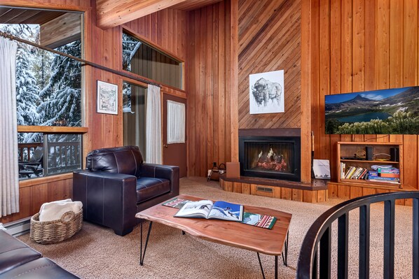 Ein gemütliches Blockhaus-Interieur mit Kamin, Ledersofa und großem Fenster mit Blick auf schneebedeckte Bäume.